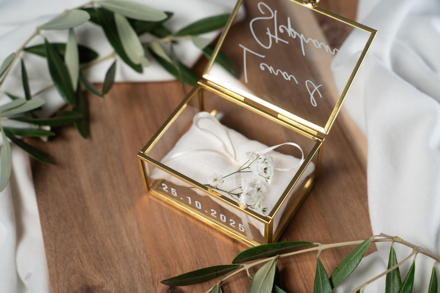 Hochwertige, personalisierbare Ringbox aus Glas mit goldfarbenem Metallrahmen, geöffnet und mit weißer Vinylschrift auf dem Deckel sowie einem aufgedruckten Hochzeitsdatum auf der Vorderseite. Im Inneren liegt ein beiges Ringkissen mit zarter Schleife, dekoriert mit kleinen weißen Blüten. Die Box befindet sich auf einer Holzoberfläche, umgeben von weichen, weißen Stoffen und Olivenzweigen im natürlichen Vintage-Stil, ideal für Hochzeit, Verlobung und Eheringe.