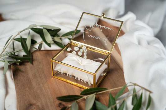 Hochwertige, personalisierbare Ringbox aus Glas mit goldfarbenem Metallrahmen, geöffnet und mit weißer Vinylschrift auf dem Deckel sowie einem aufgedruckten Hochzeitsdatum auf der Vorderseite. Im Inneren liegt ein beiges Ringkissen mit zarter Schleife, dekoriert mit kleinen weißen Blüten. Die Box befindet sich auf einer Holzoberfläche, umgeben von weichen, weißen Stoffen und Olivenzweigen im natürlichen Vintage-Stil, ideal für Hochzeit, Verlobung und Eheringe.